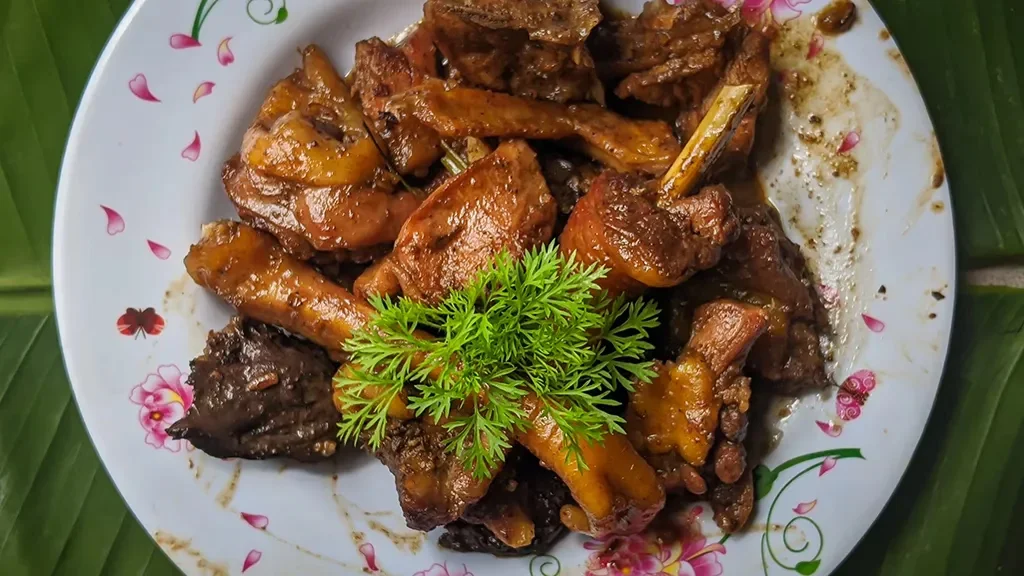 A plate of Filipino Adobo sa Gata, chicken pieces braised in vinegar, garlic, and coconut milk until deeply glazed, garnished with fresh green herbs and served on a floral-rimmed white plate atop a banana leaf.