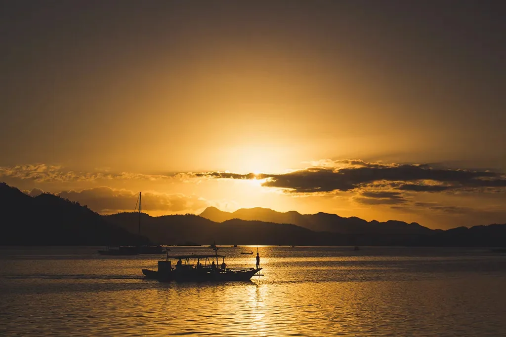 A silhouetted bangka boat with a figure standing at the bow crosses the golden waters of Coron Bay at sunset in Palawan, Philippines, with the mountain ridgeline of Busuanga Island and dramatic clouds glowing amber and orange against the evening sky.