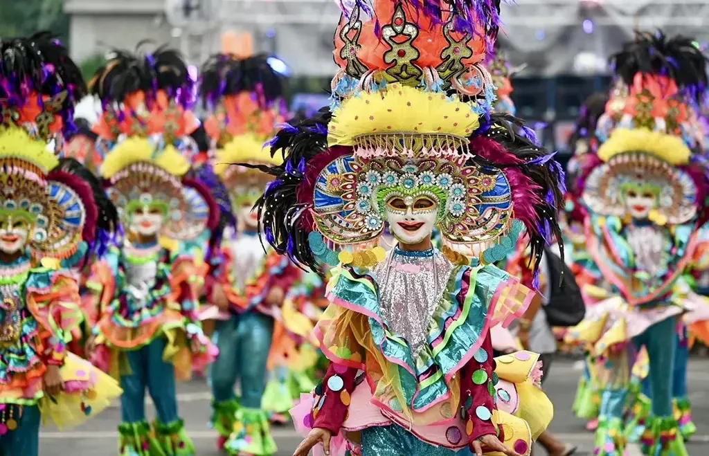 A close-up of a Sinulog Festival parade dancer in Cebu City, Philippines, wearing an ornate jeweled mask, towering feathered headdress, and a multicolored ruffled sequined costume, with a line of similarly costumed dancers visible in the background.