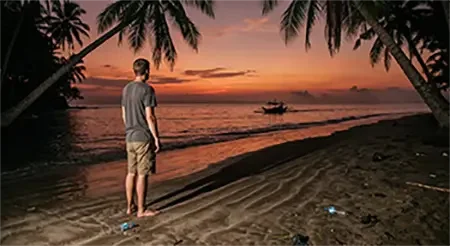 Stranded traveler watching a Bangka motor away from a remote Philippine island beach at sunset, island hopping in the Philippines gone wrong